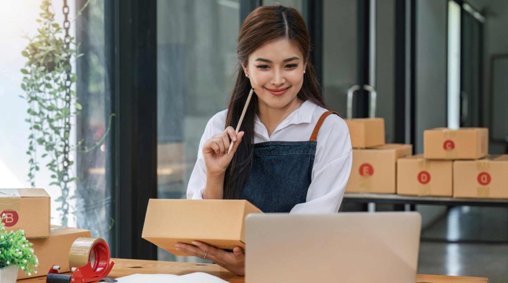 Woman sitting at a table holding a cardboard box, smiling at a laptop. Packing materials, tape, and boxes are on the table. She appears to be working in a small business or online store setting.