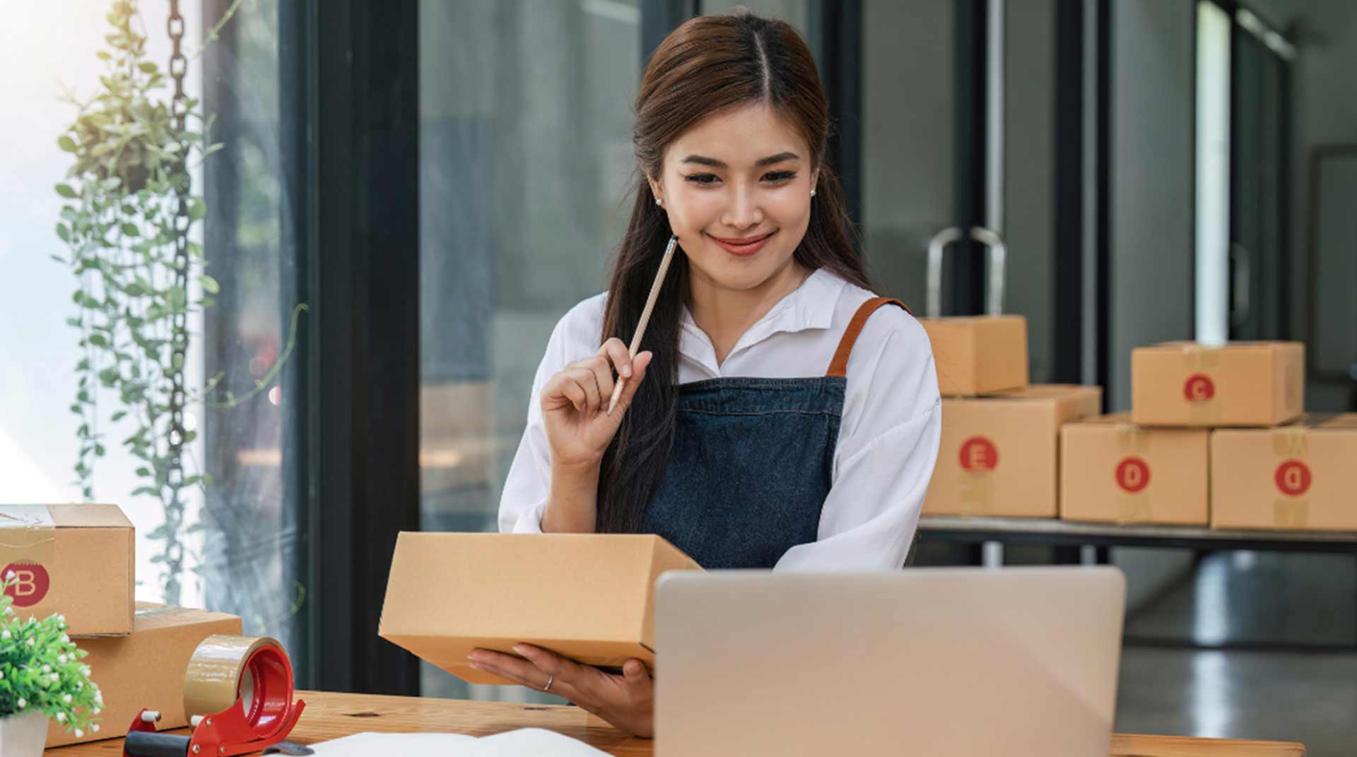 Woman sitting at a table holding a cardboard box, smiling at a laptop. Packing materials, tape, and boxes are on the table. She appears to be working in a small business or online store setting.