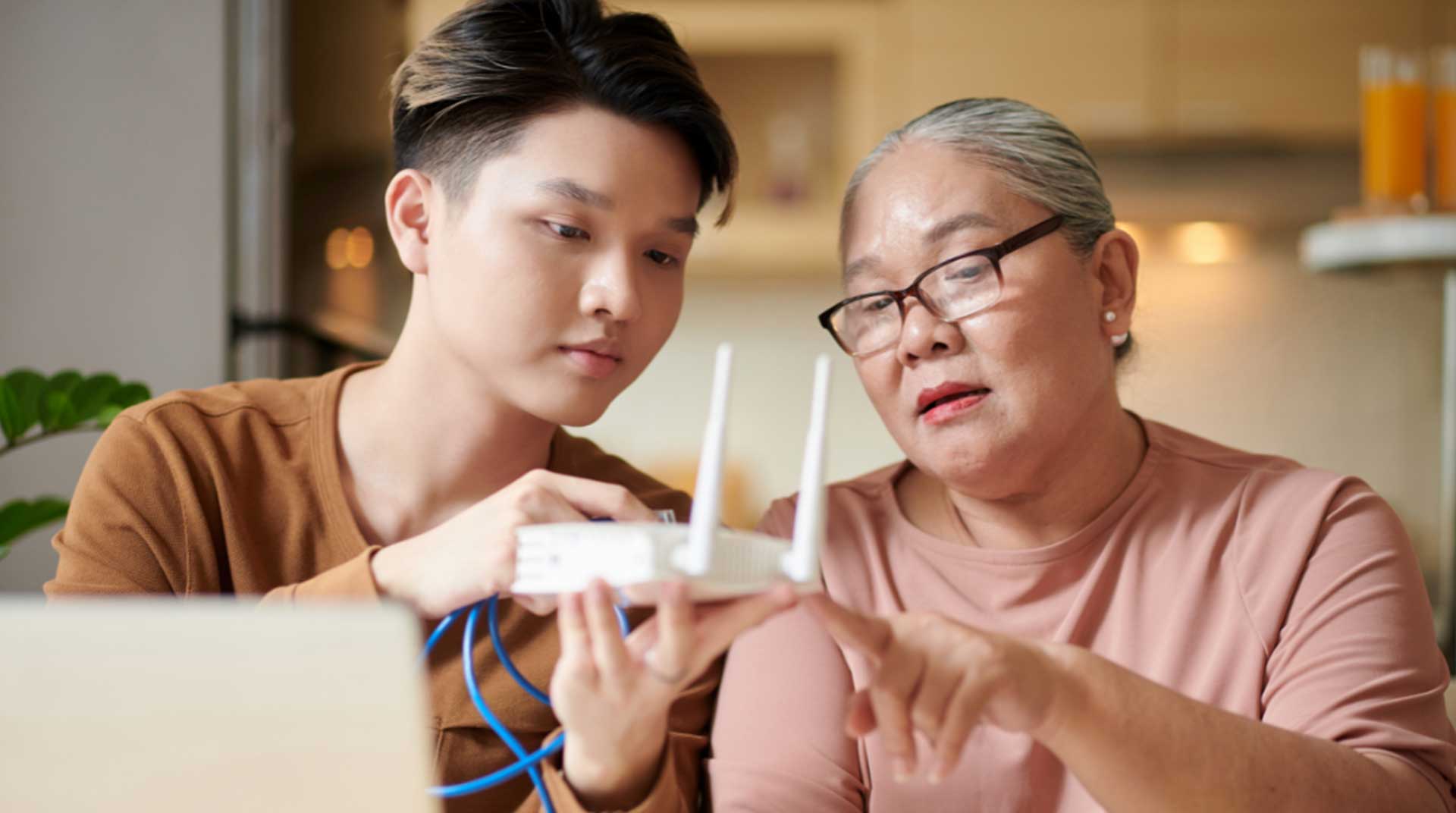 A young man and an older woman are sitting together at a table, examining a Wi-Fi router. The man holds the device, while the woman points at it. They appear focused, indoors with a laptop and kitchen items visible in the background.