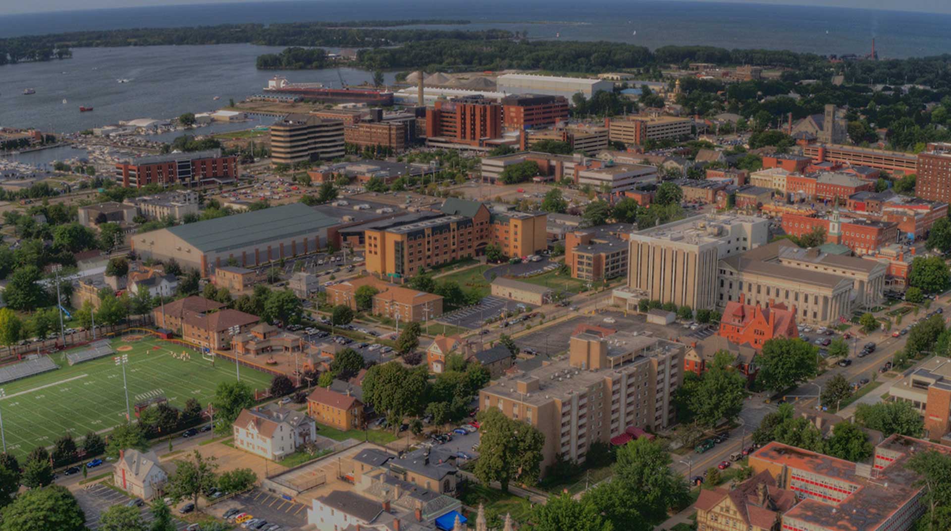 Aerial view of Erie, Pennsylvania with Fiber Internet, showcasing modern and historical buildings, a sports field in the foreground, tree-lined streets, and a bustling harbor with boats. The backdrop reveals a vast body of water and distant land on the horizon.
