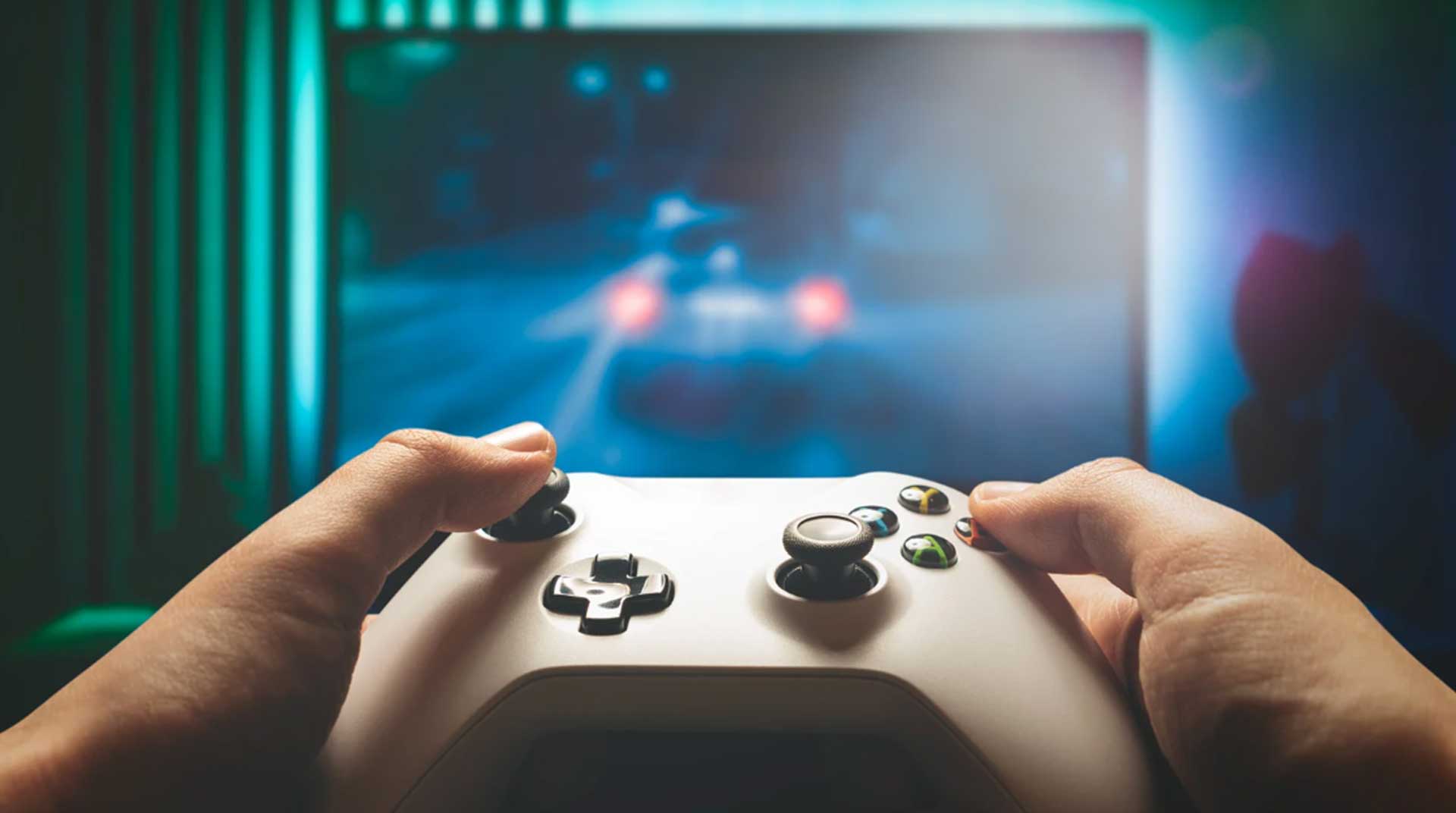 Hands holding a white video game controller in front of a blurry, illuminated screen displaying a racing game. The room has a dark ambiance with a green and blue glow.