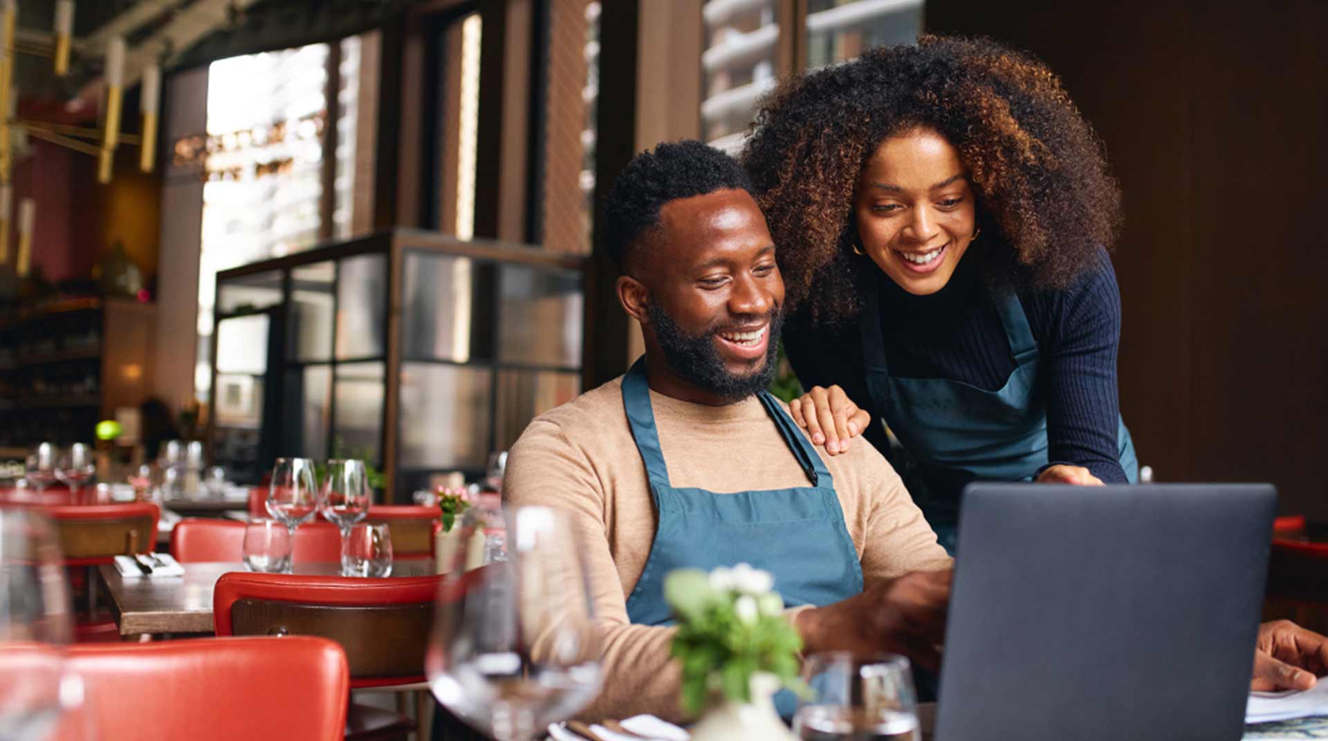 A man and woman, both wearing aprons, stand behind a laptop in a cozy restaurant setting. They are smiling and appear engaged with the screen. The table is set with glasses and a small plant centerpiece.
