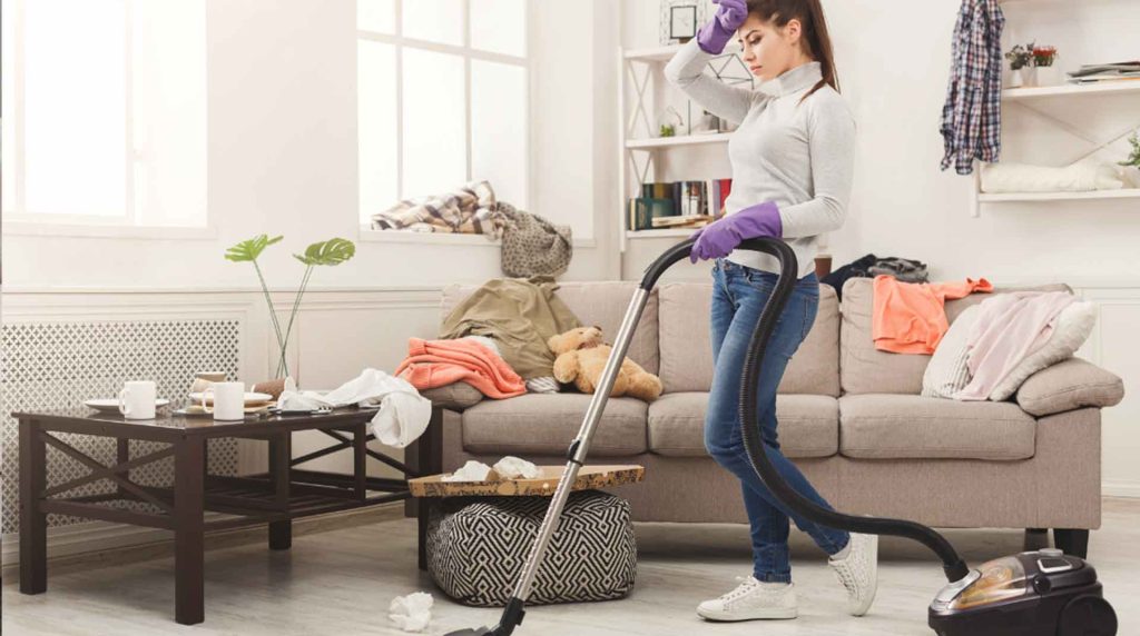 A woman wearing a gray sweater and purple gloves vacuums a cluttered living room. The couch is covered with clothes and a teddy bear. A coffee table is cluttered with cups and tissues. She wipes her forehead, appearing tired.