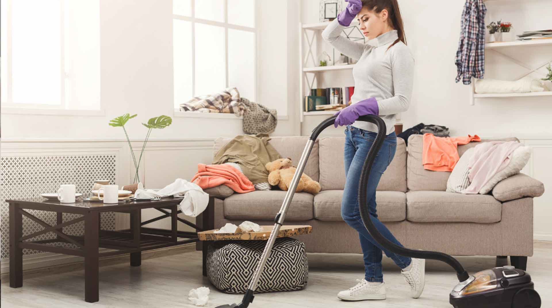 A woman wearing a gray sweater and purple gloves vacuums a cluttered living room. The couch is covered with clothes and a teddy bear. A coffee table is cluttered with cups and tissues. She wipes her forehead, appearing tired.