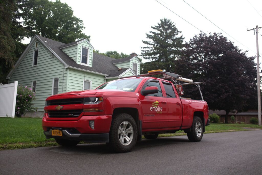 A red Empire-branded pickup truck with ladders on top is parked on a residential street in front of a light green house with white trim and a sloped roof, possibly there to install or fix streaming TV services. Trees and grass surround the home.
