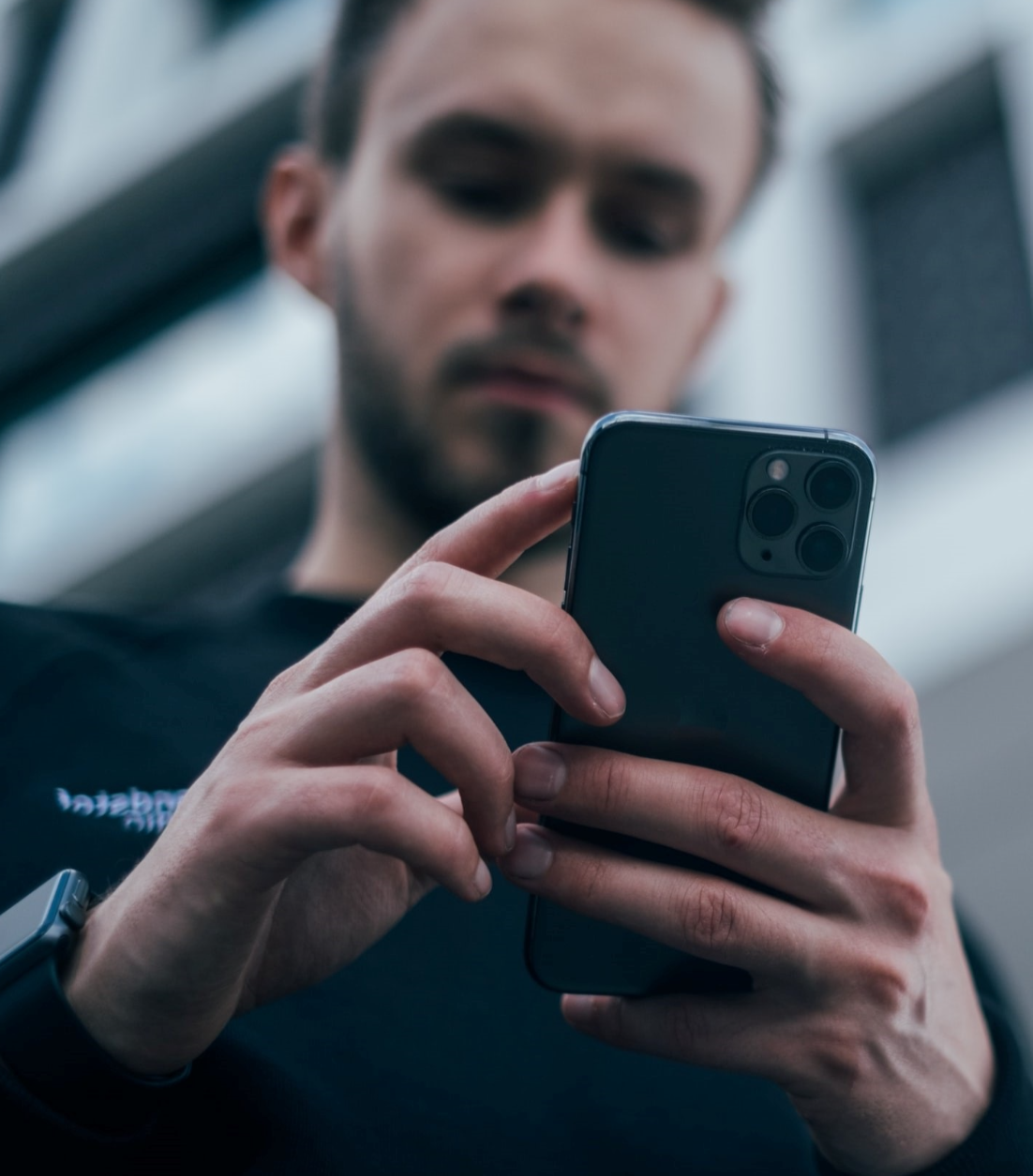 A person wearing a dark shirt and smartwatch is holding and using a smartphone, with the photo focused on their hands and phone, and their face slightly blurred in the background.