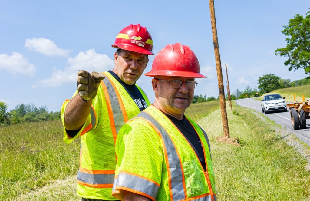 Two construction workers in yellow vests and red hard hats stand beside a rural road. One points toward the camera while the other looks on, as if discussing Streaming TV. A white car and roadwork equipment are visible in the background.