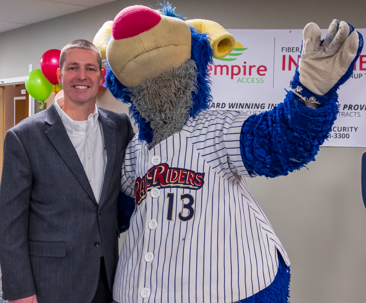 A man in a suit stands smiling next to a person in a blue and yellow bear mascot costume wearing a pinstriped jersey with the number 13. They are indoors, with colorful balloons and a business sign in the background.