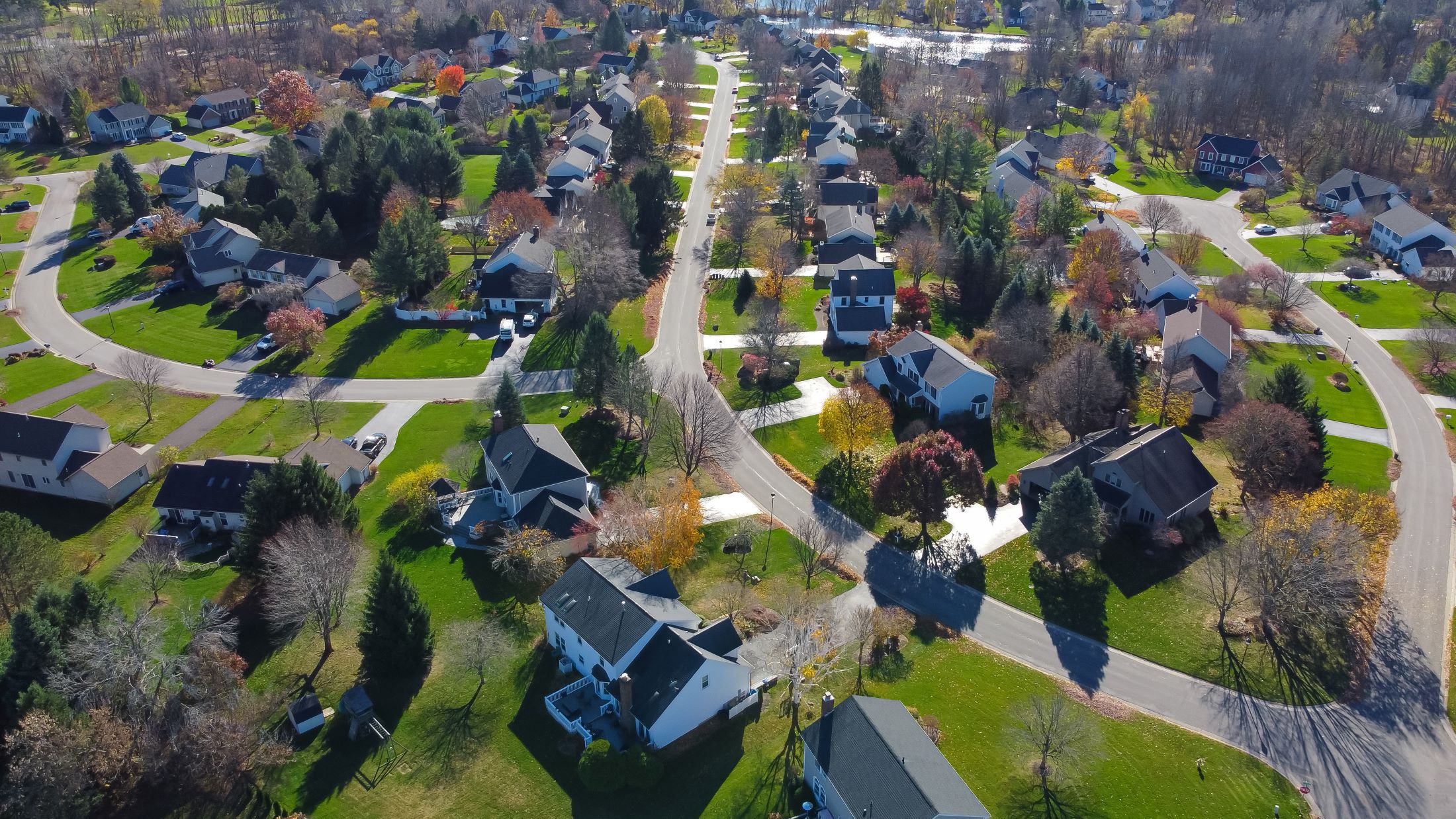 Aerial view of a suburban neighborhood with curving streets, green lawns, and houses surrounded by trees with autumn foliage. The scene is bright and peaceful under clear skies.