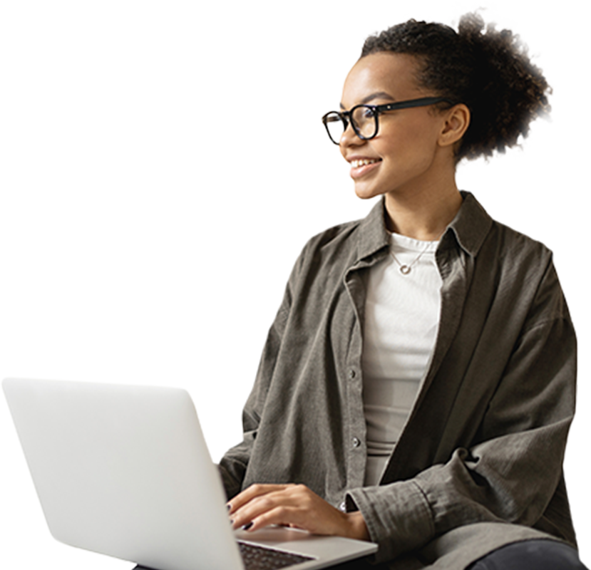 A person with curly hair and glasses is smiling while using a laptop. They are wearing a gray button-up shirt over a white top and sitting against a plain background.