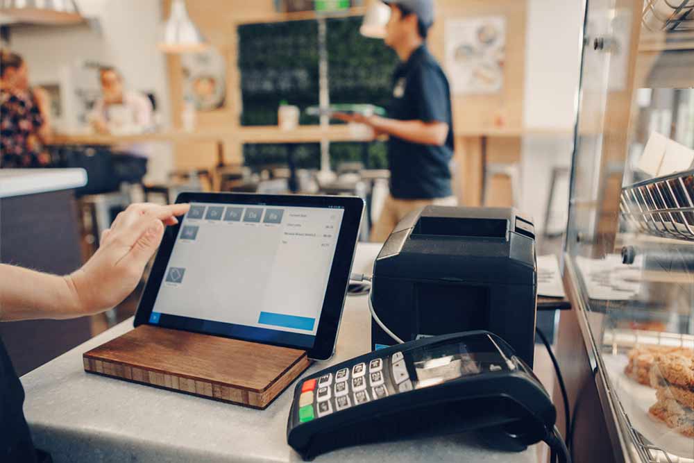 A person uses a touchscreen tablet at a café counter, next to a payment terminal and receipt printer—demonstrating how Dark Fiber for Businesses can support seamless, high-speed transactions as a staff member in uniform walks by in the background.