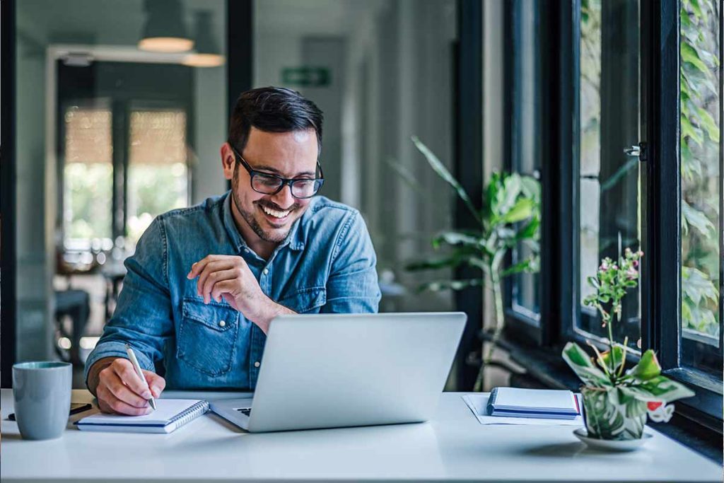 A smiling man wearing glasses and a denim shirt sits at a desk by a window, working on a laptop with notes in hand. Surrounded by plants and natural light, he researches Multi-Dwelling Internet Services in his modern workspace.
