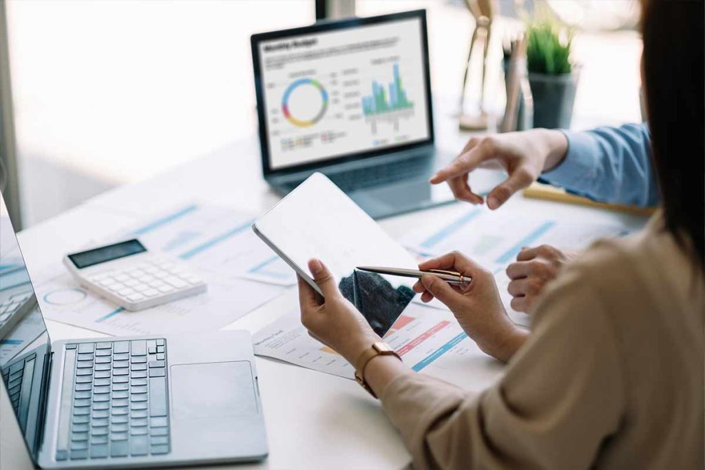 Two people are discussing financial charts and graphs related to Multi-Dwelling Internet Services at a desk with a laptop, tablet, papers, and a calculator. One points at the tablet while the other gestures as data and colorful graphs appear on a screen.
