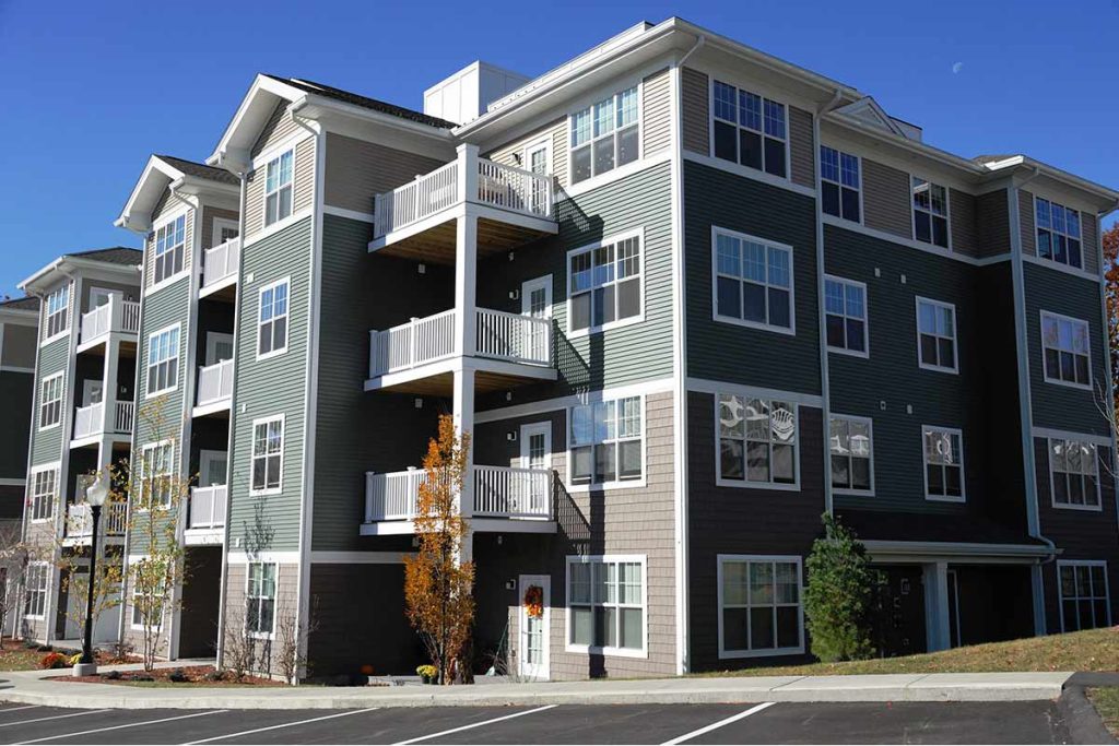 A modern, multi-story apartment building with green and beige siding, white trim, and several balconies. The property is ideal for Multi-Dwelling Internet Services, with a landscaped area and an empty parking lot under a clear blue sky.