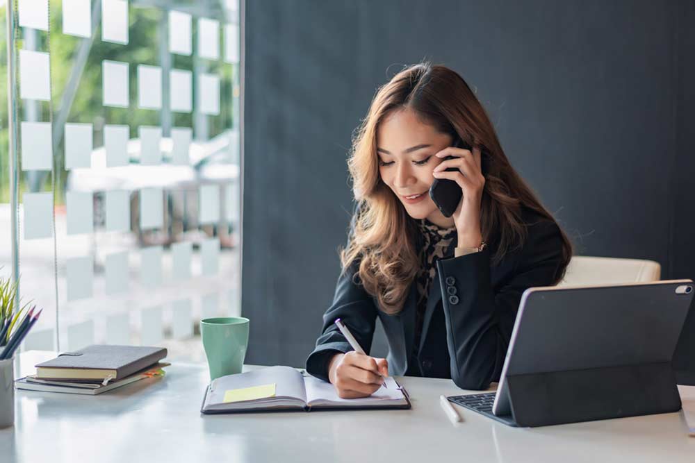 A woman in a black blazer sits at a desk by a window, talking on a phone while writing in a notebook. An open laptop, green mug, and notebooks are on the desk. She appears focused and engaged.