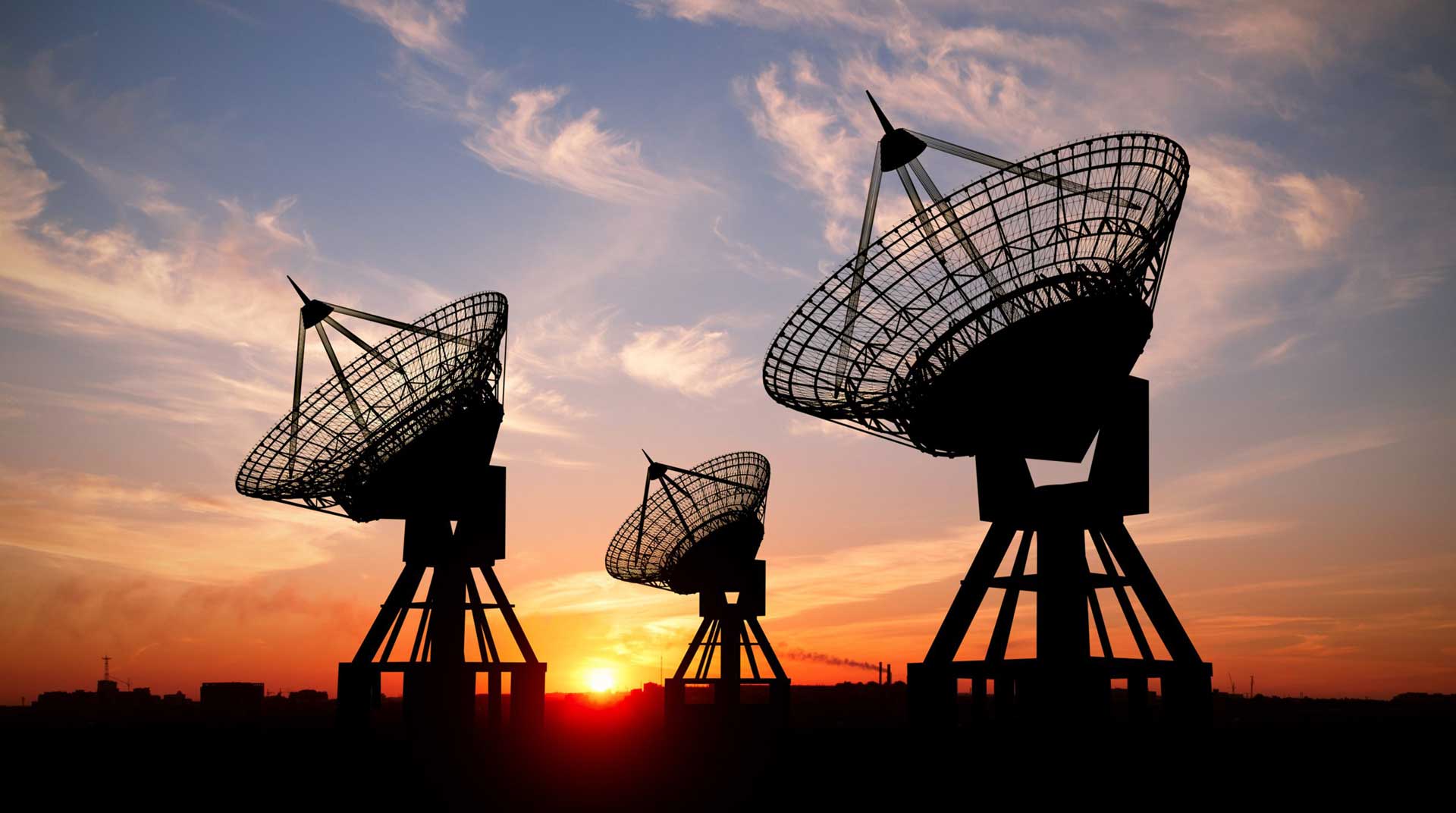 Three large solar satellite dishes are silhouetted against a colorful sunset sky with scattered clouds. The sun sets on the horizon, casting a warm glow behind the structures.