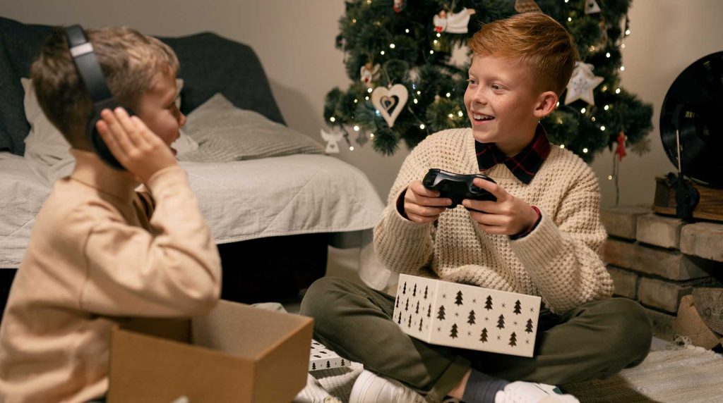 two boys sitting on the floor next to a Christmas tree looking at their new gifts, a video game system and wireless headphones
