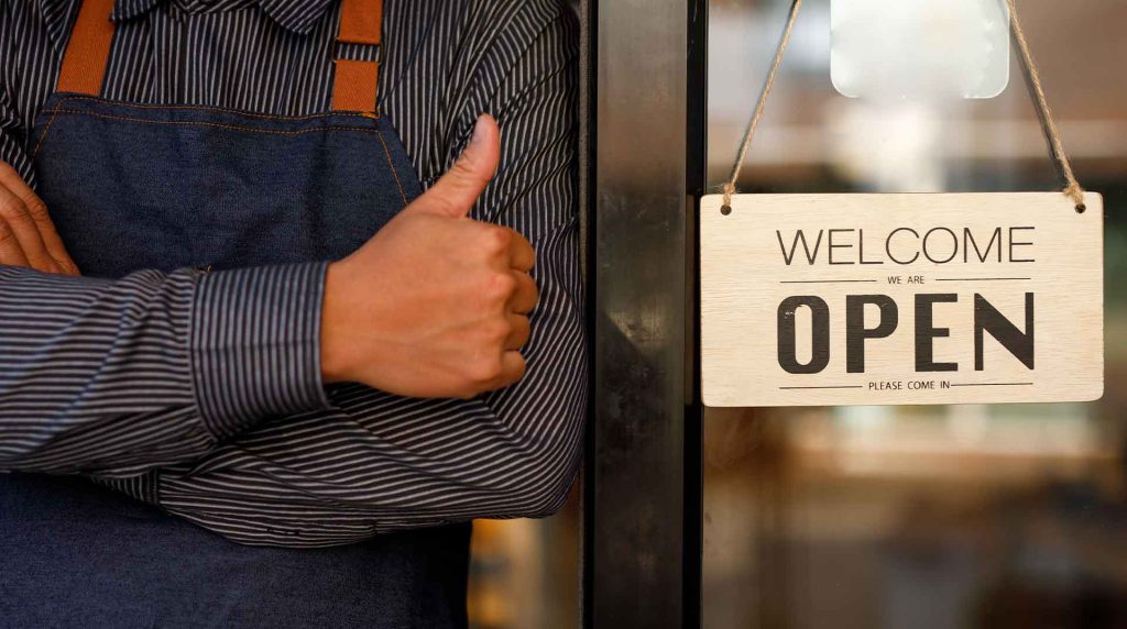 close up of business owner standing by the front door with a "Welcome - We're Open" sign giving a thumbs up to business internet solutions
