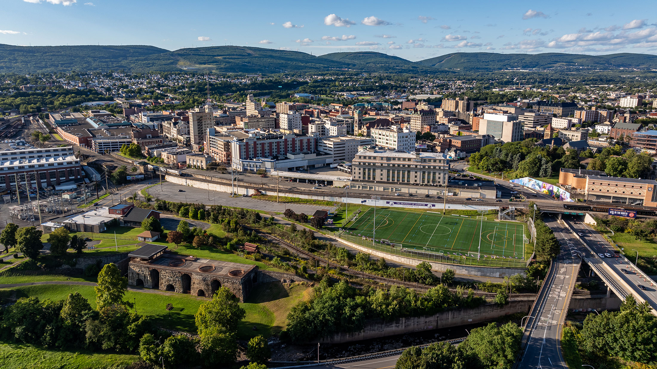 Aerial view of a cityscape featuring a soccer field in the foreground surrounded by roads and trees. Numerous buildings, including some taller structures, are visible in the background with green hills under a partly cloudy sky.
